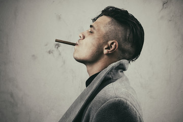 Young man in grey coat smoking cigar on the dirty background with vignette. Photo of smoking guy...