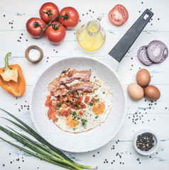 homemade scrambled eggs with bacon, tomatoes, green onions, bell pepper and seasonings on a white frying pan, around lie the ingredients top view