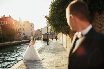 Look over groom's shoulder at stunning bride standing before the canal in Venice