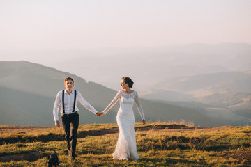 Cheerful newlyweds walk with black puppy on a hill before beautiful landscape