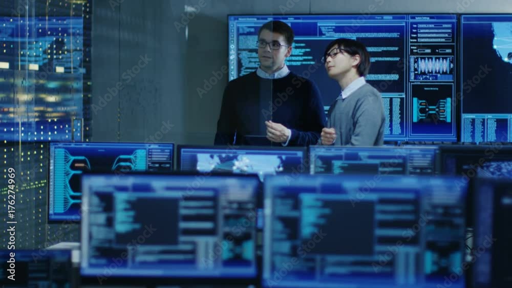 Poster In the System Control Room Project Manage and IT Engineer Have Discussion, they're surrounded by Multiple Monitors with Graphics. They Work in a Data Center on Data Mining, AI and Neural Networking.