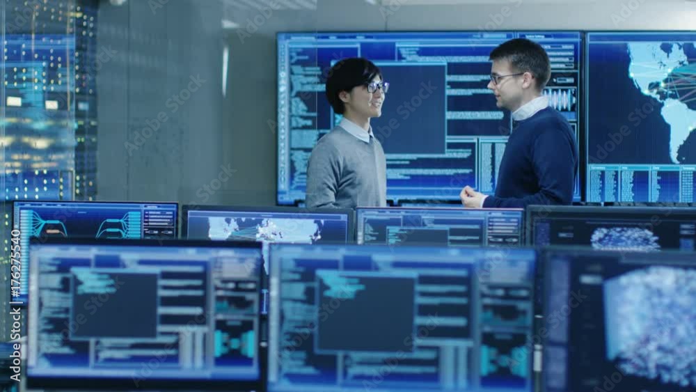 Poster In the System Control Room Project Manage and IT Engineer Have Discussion, they're surrounded by Multiple Monitors with Graphics. They Work in a Data Center on Data Mining, AI and Neural Networking.