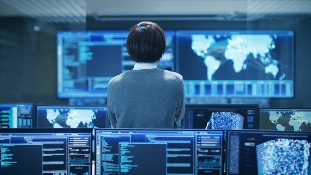 In the System Control Room Technical Operator Stands and Monitors Various Activities Showing on Multiple Displays with Graphics. Artificial Intelligence, Big Data Mining, Neural Network.