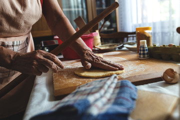 Close up of senior female baker hands kneading dough in kitchen. She is using rolling pin.