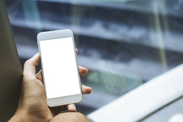 Mockup image of a man's hand holding white mobile phone with blank screen on thigh with white canvas shoes in modern cafe with feeling relax