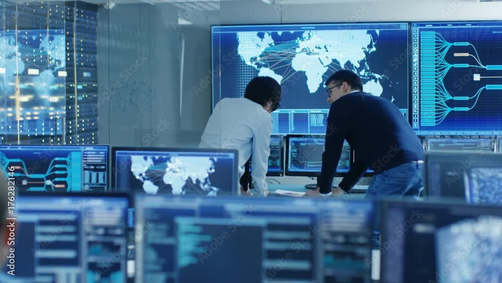 Poster In the System Control Room Project Manage and IT Engineer Have Discussion, they're surrounded by Multiple Monitors with Graphics. Big Monitor Shows Interactive Logistics Map. 