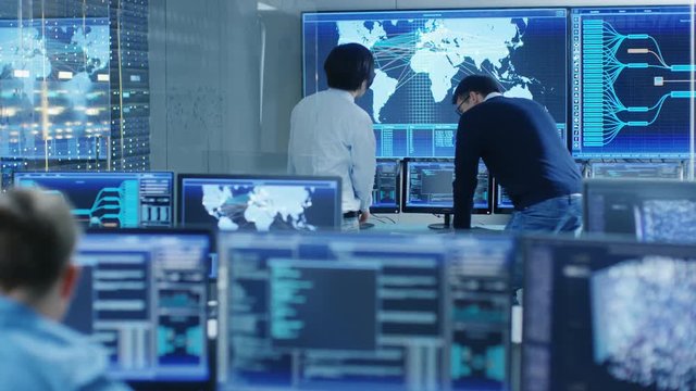 In the System Control Room Project Manage and IT Engineer Have Discussion, they're surrounded by Multiple Monitors with Graphics. Big Monitor Shows Interactive Logistics Map. 