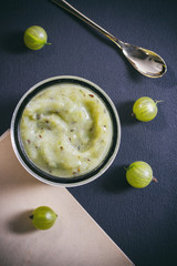 Gooseberry jam, golden spoon and baking paper on dark, view from top