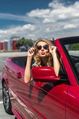 Portrait of beautiful fashion woman model in sunglasses sitting in luxury red convertible car with sea and sky background. Young woman driving on road trip on sunny summer day. Sea and sky. 
