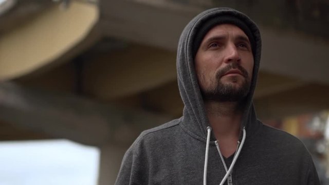 thoughtful bearded man wearing hat and grey hoodie standing under footbridge dreaming looking on camera in slow motion 