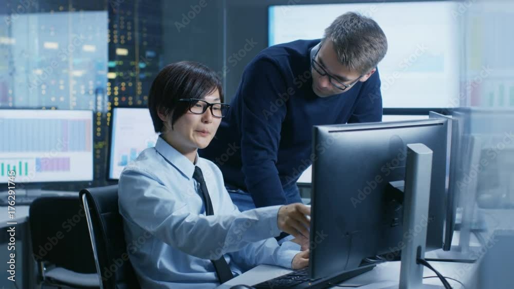 Fototapete In the Data Mining Center Chief Statistician Consults His Young Assistant sits at His Workstation Surrounded with Monitors Displaying Data and Graphs. Shot on RED EPIC-W 8K Helium Cinema Camera.