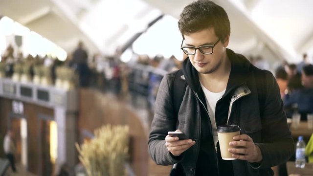 Handsome young man wearing glasses and a sweatshirt is texting and drinking a coffee to go at a farmer s market. Handheld real time medium shot