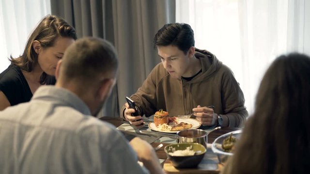 Family eating dinner at home, son looking at smartphone
