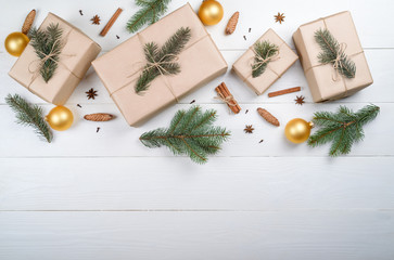 Christmas background with gift boxes wrapped in kraft paper, fir tree branches, golden glass balls, pine cones, cinnamon and stars anise on white wooden background, free space. Flat lay, top view