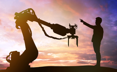 Industry 4.0 technology , artificial intelligence trend concept. Silhouette of business man point finger forward to heavy automation robot arm machine. Vivid twilight sunset sky background.