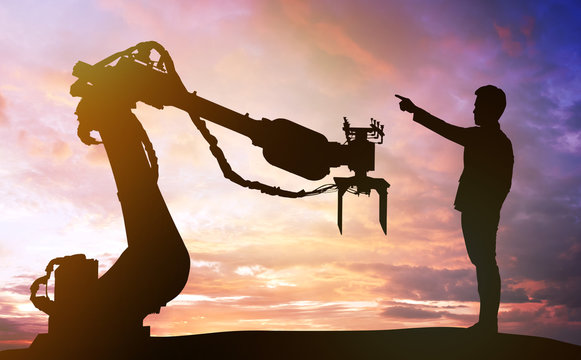 Industry 4.0 Technology , Artificial Intelligence Trend Concept. Silhouette Of Business Man Point Finger Forward To Heavy Automation Robot Arm Machine. Vivid Twilight Sunset Sky Background.