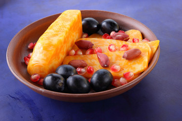 Orange cheese, sliced cheese, a sort of hard cheese, marbled cheese, dark blue grapes, peanuts and pomegranate seeds in a clay plate on a dark blue background