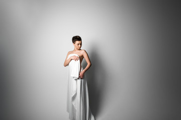 Sensual naked young adult caucasian woman, wrapped in a satin, silk sheet against grey background. High contrast