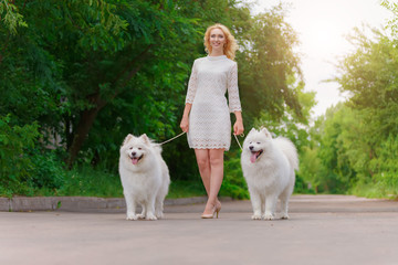 Beautiful young blond girl in dress walking with two white fluffy dogs in summer garden