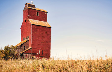 One abandoned old red wooden grain storage elevator behind tall weeds in autumn landscape