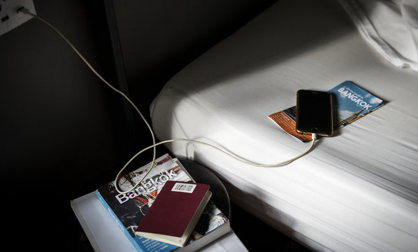 A Mobile Phone Charging Lying On The Bed With A Bangkok, Thailand Travel Guide Book And Brochure