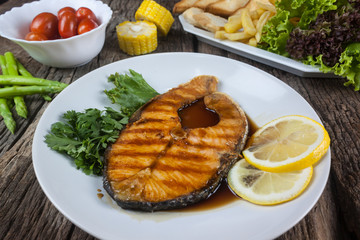Salmon steak with sauce on a white plate with lemon on plate and crispy French fries. Many vegetables are placed around the dish on the wooden floor.