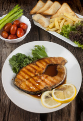 Salmon steak with sauce on a white plate with lemon on plate and crispy French fries. Many vegetables are placed around the dish on the wooden floor.
