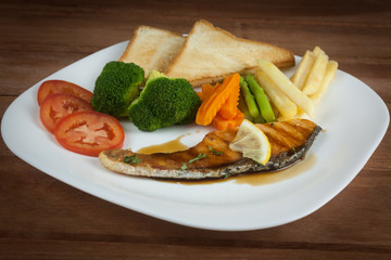 Grilled salmon steak slices until cooked, placed on a white plate with lemon on fish and vegetables, placed around the plate on a white wooden floor.