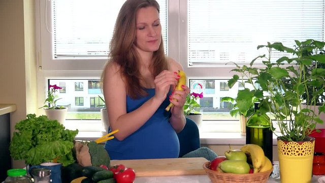 Hungry smiling pregnant woman with big belly peel off banana in kitchen