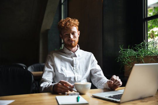 Photo of young smiling readhear bearded man holding coffe cup, looking  at camera while working at cafeteria
