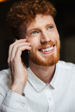 Close-up portrait of handsome smiling readhead bearded man in white shirt tolking on mobile phone