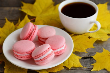 Obraz na płótnie Canvas Sweet French macaroons. Light pink macaroons on a white plate, a cup of coffee, yellow leaves on an old wooden background. Autumnal breakfast concept. Closeup