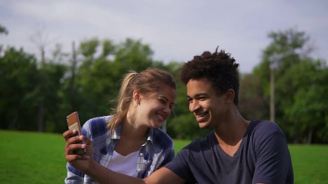 Cute friends resting in a park, sitting on the grass, looking at the smartphone and laughing. Mixed race couple sitting on the grass and selects pictures in their smartphone