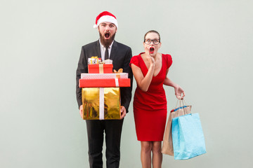 No way! Amazed bearded man in red hat and woman in red dress, holding christmas gifts and colorful packages and looking at camera with wonder face. Studio shot on gray background