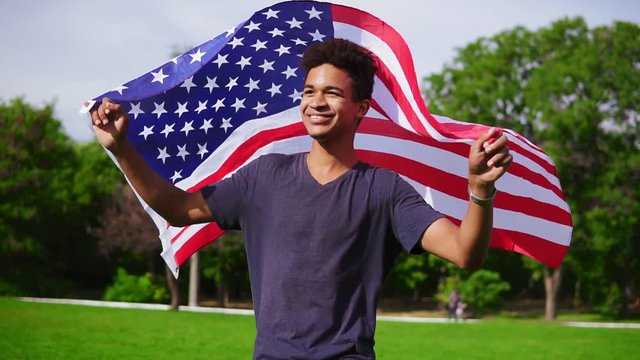 Attractive african american man holding American flag in his hands on the back walking in the green field and smiling proudly. Patriotic concept