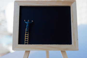 Miniature people: small figurine character over ladder in front of a blackboard with copyspace