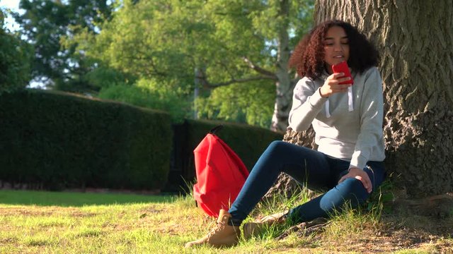 4K video clip of beautiful happy mixed race African American girl teenager leaning against a tree with a red backpack and using a cell phone camera to take pictures and social media
