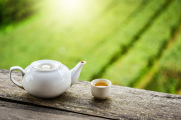 White teapot and teacup on wooden deck with green tea plantation garden background. Soft light effect added. Fresh green tea is very healthy.