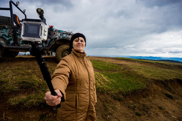 Senior woman in mountains taking selfie on action camera with dramatic sky and tourist jeep on background in autumn day