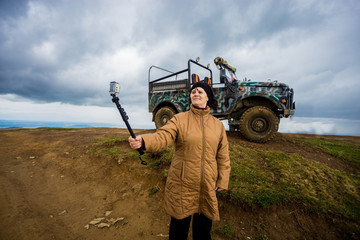 Senior woman in mountains taking selfie on action camera with dramatic sky and tourist jeep on...