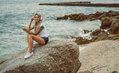 Beautiful blonde woman sitting on a rock by the sea and listening to music on stylish headphones on the tablet. hands with a tattoo. The style of summer holiday. Modern girl