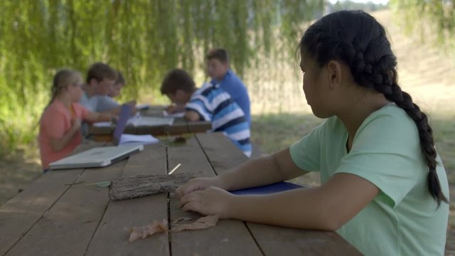 Lonely girl sits alone looking sad at wooden table during an outdoor class at summer camp with other students in background. 4K