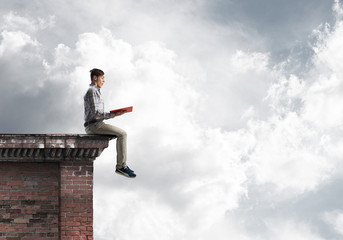 Man on roof edge reading book and cloudscape at background