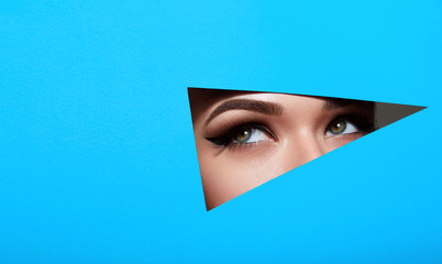A girl with beautiful green bright eyes with brown shadows and expressive eyebrows looks into the hole of colored paper.Fashion, beauty, make-up, cosmetics, make-up artist, beauty salon,business card. - obrazy, fototapety, plakaty