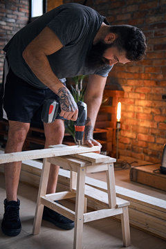 Carpenter drilling a hole in a board in a room with loft interior.