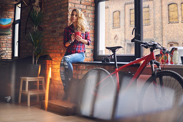 Blond female drinks hot coffee near the window.