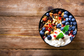 Tasty homemade granola, yogurt, fresh organic berries, pomegranate, mint on rustic wooden background with copy space, top view.