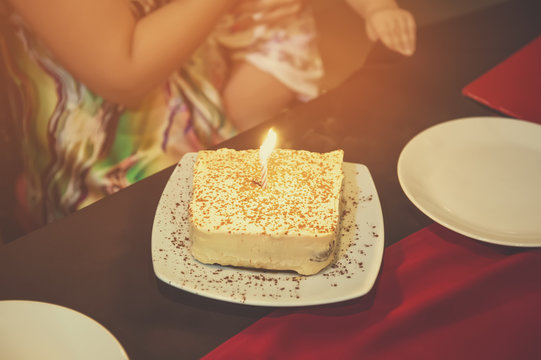 Piece of cake with one burning candle on a white plate on the table in the cafe. Birthday.