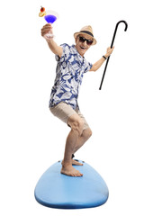 Overjoyed senior with a cocktail and a cane standing on a surfboard