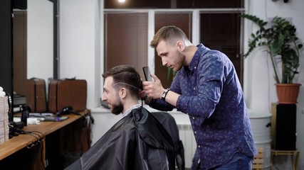 Making haircut. Young bearded man getting haircut by barber while sitting in chair at barbershop